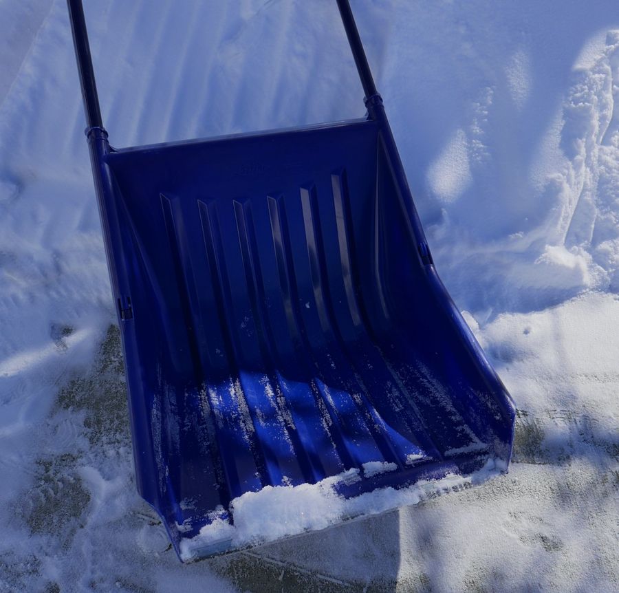 A large blue plastic shovel leaning on a snow pile on a driveway. There is snow on the metal edge of the shovel. There are lines on the snow pile where the shovel was pushed up atop of it.